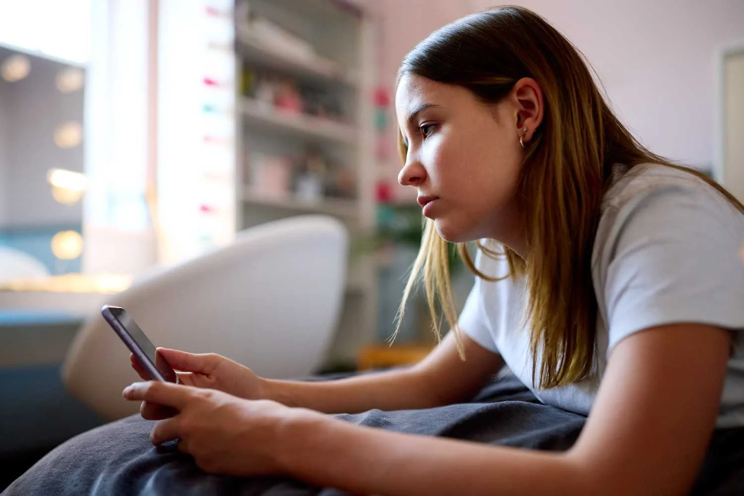 Pensive teen girl in t-shirt on bed with phone in sunlit room, depicting original 2026 teen mental health statistics: 47% device worry, 68% untreated moods.