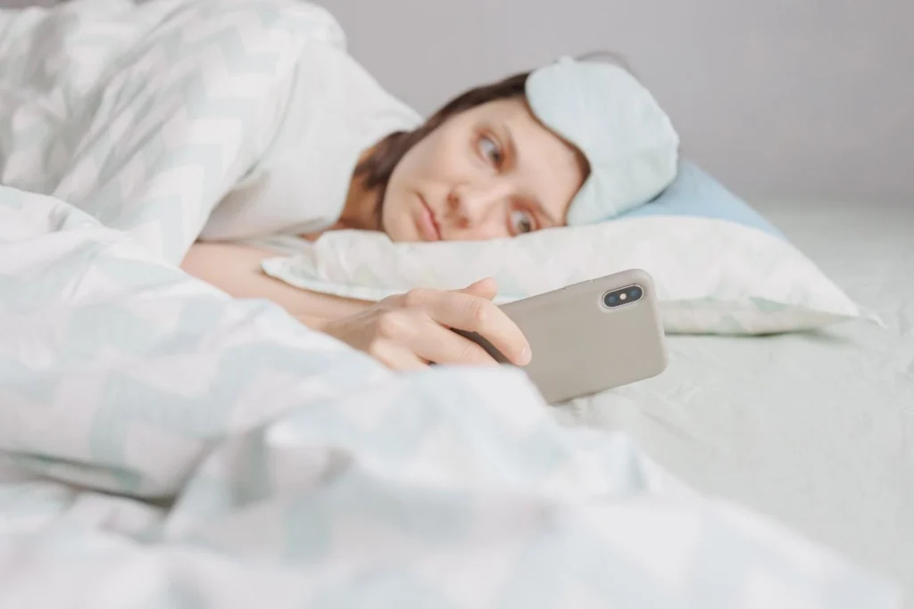 Woman lying in bed with a sleep mask pushed up, holding a smartphone, unable to sleep — representing insomnia disorder and how online insomnia treatment works