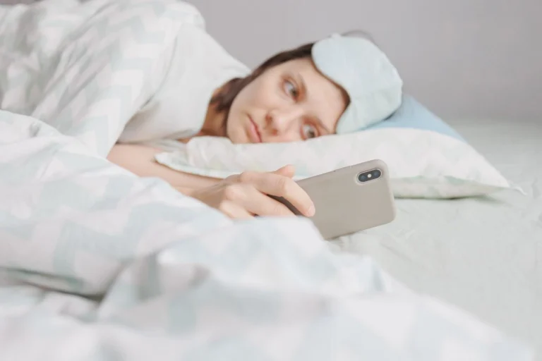 Woman lying in bed with a sleep mask pushed up, holding a smartphone, unable to sleep — representing insomnia disorder and how online insomnia treatment works